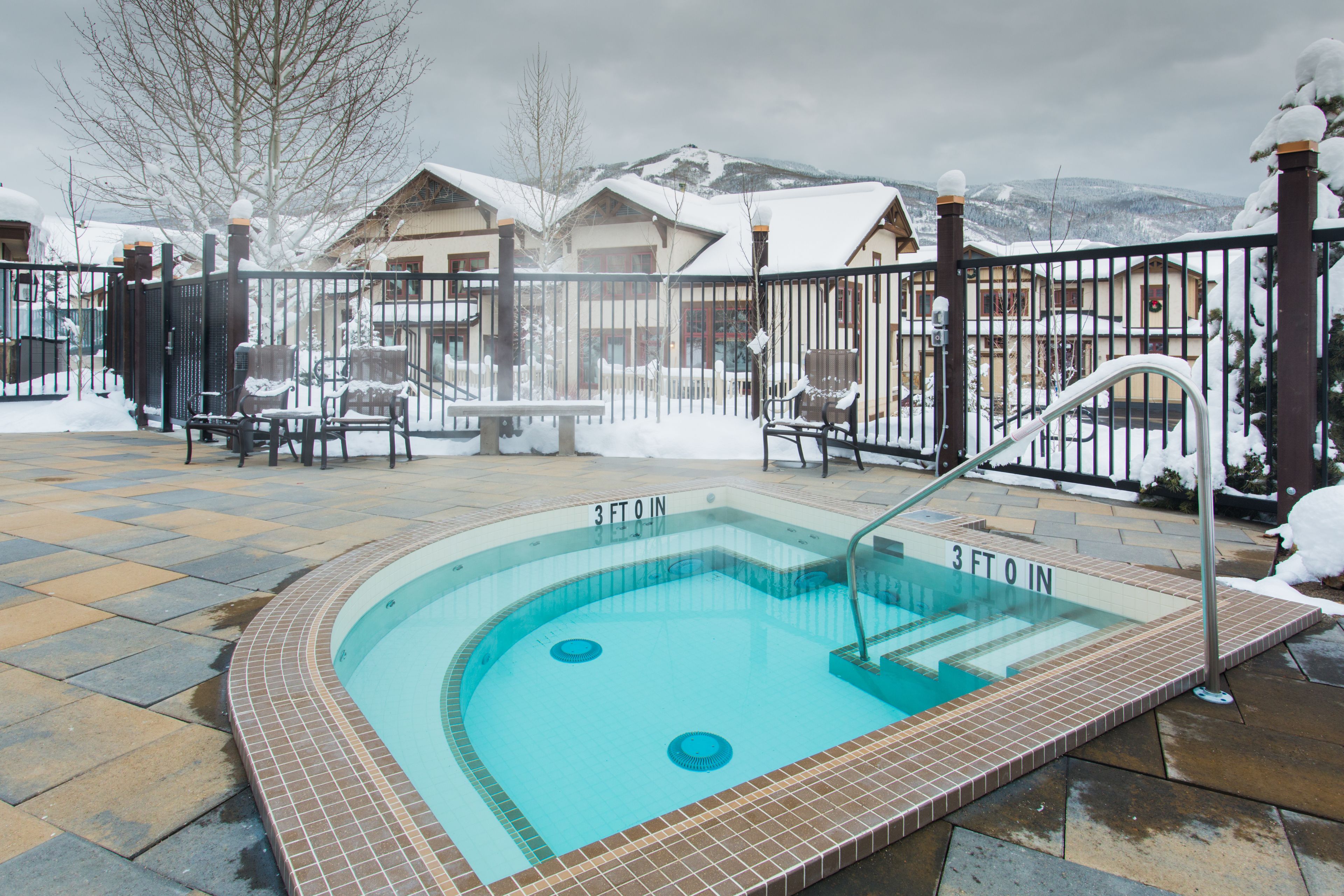 A 3-foot pool with a tiled edge, surrounded by a fence, with snow-covered buildings and mountains in the background.