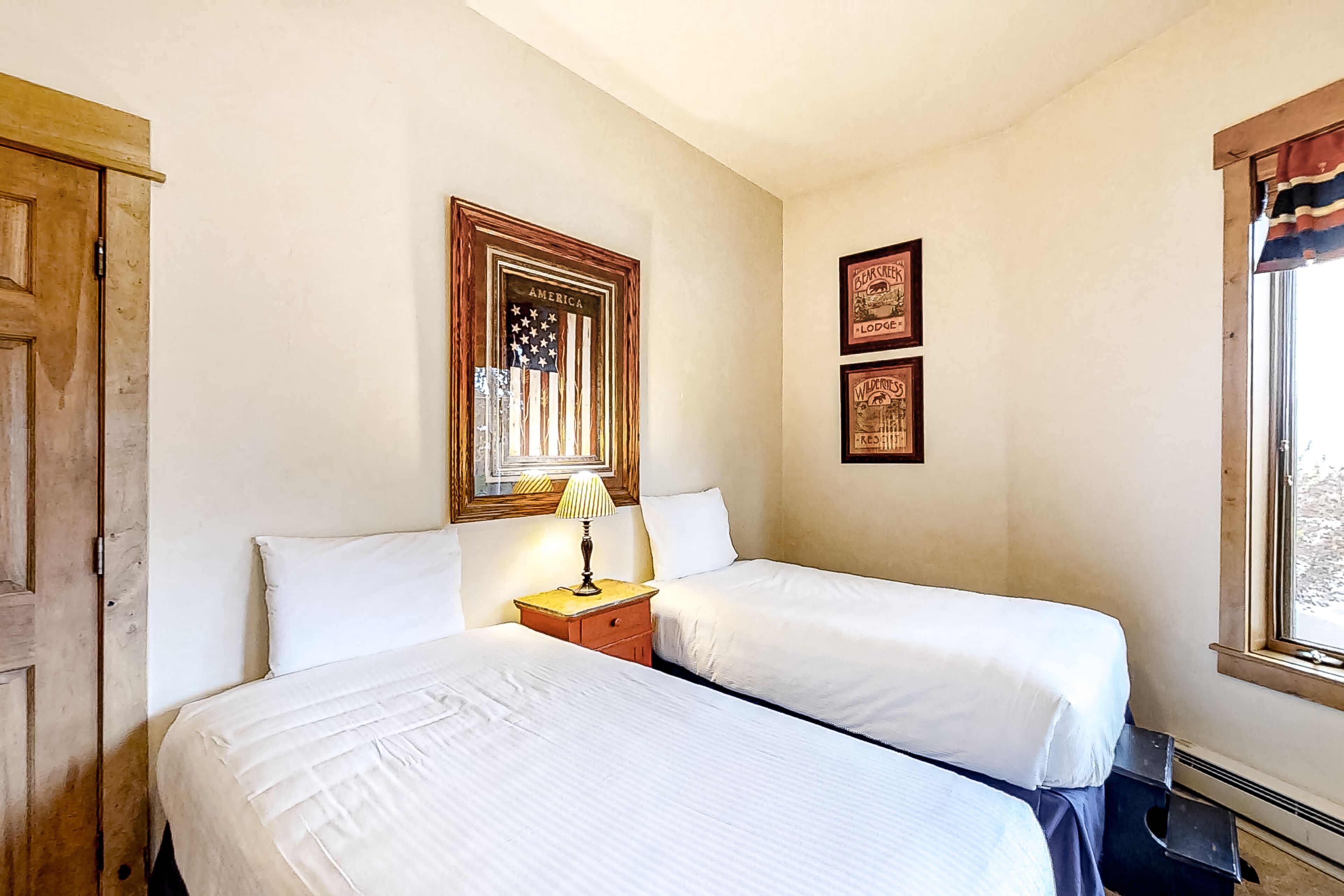 A hotel room with two beds, a wooden door, a framed American flag, and two framed pictures on the wall.