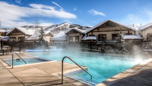 Un espace piscine avec un jacuzzi, entouré de bâtiments aux toits enneigés et dominé par un paysage montagneux.