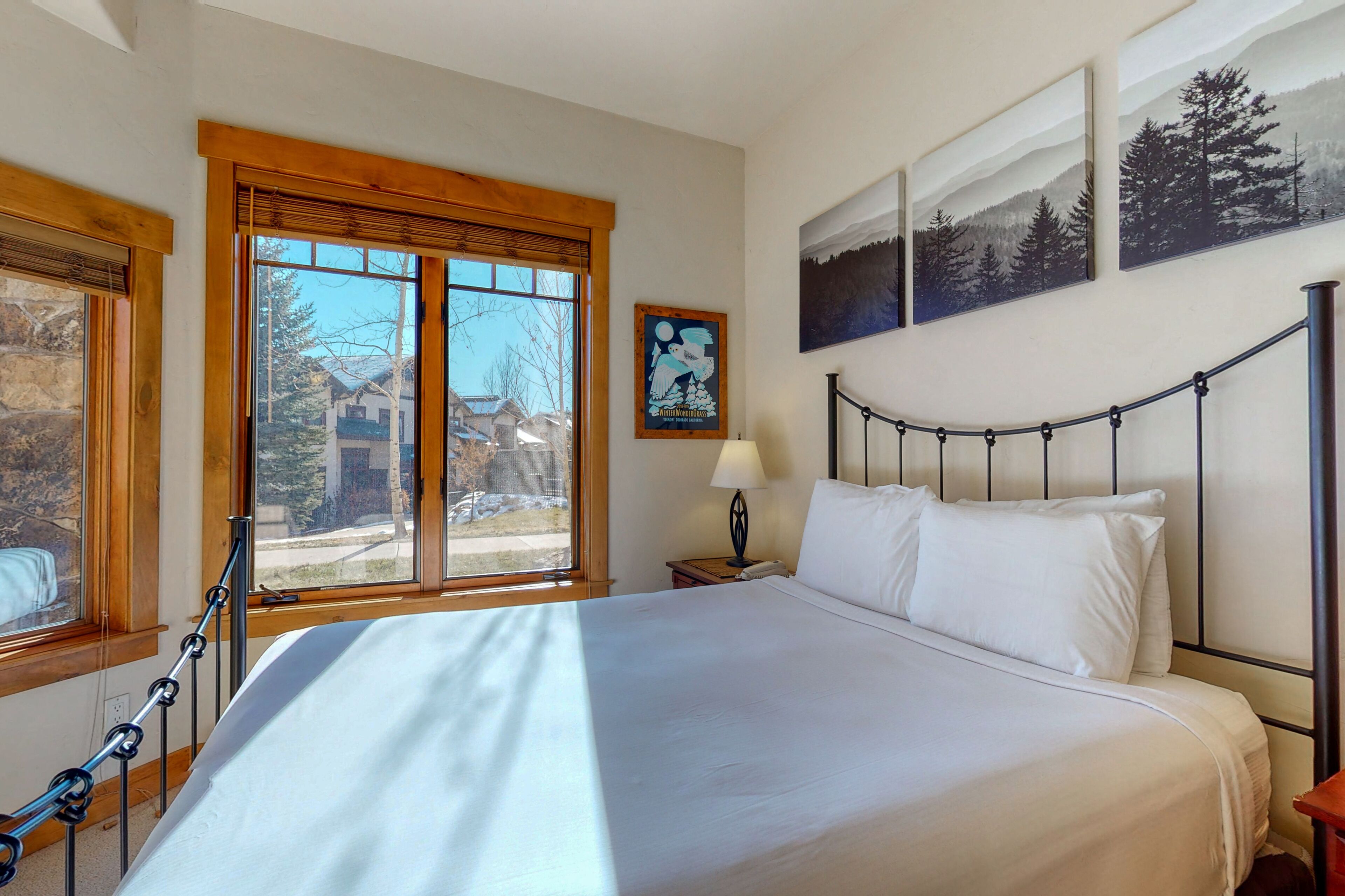 Une chambre avec un grand lit, une fenêtre donnant sur des maisons, une tête de lit en bois et un mur orné de trois tableaux encadrés.