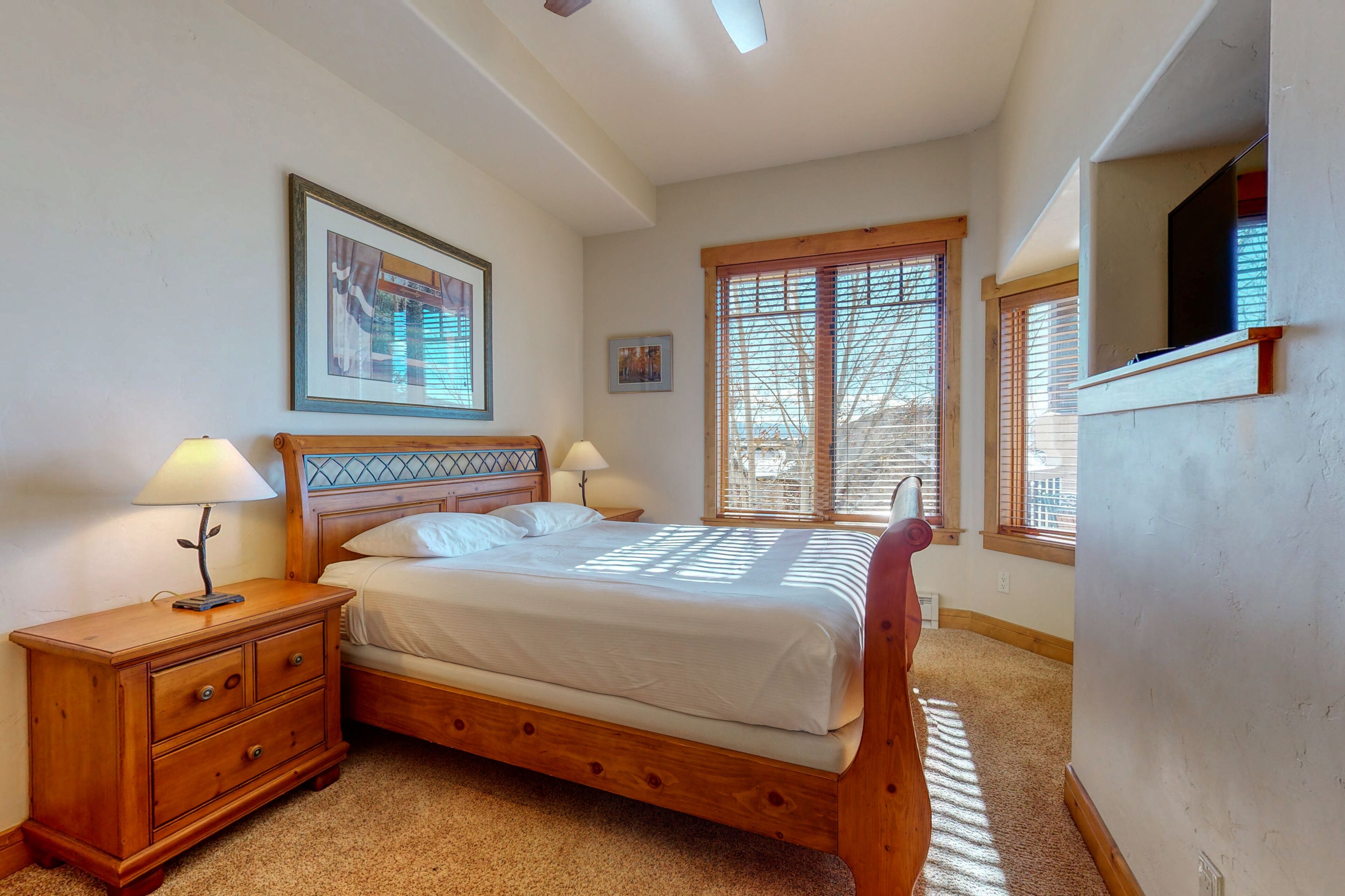 A bedroom with a wooden bed, two bedside tables with lamps, a framed picture on the wall, and a window with blinds.
