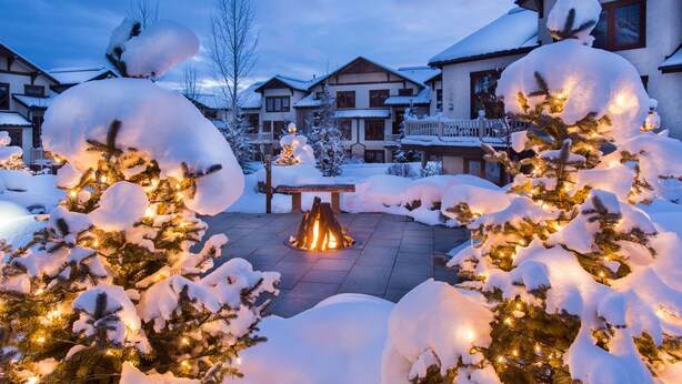 Un espace extérieur enneigé avec un foyer, des arbres couverts de neige et un bâtiment en arrière-plan.