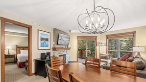 A dining area with a wooden table, chairs, and a chandelier. There is a fireplace with a stone surround and a television mounted on the wall. The room has large windows with a view of trees. - Horstman House by Whistler Premier (Whistler)