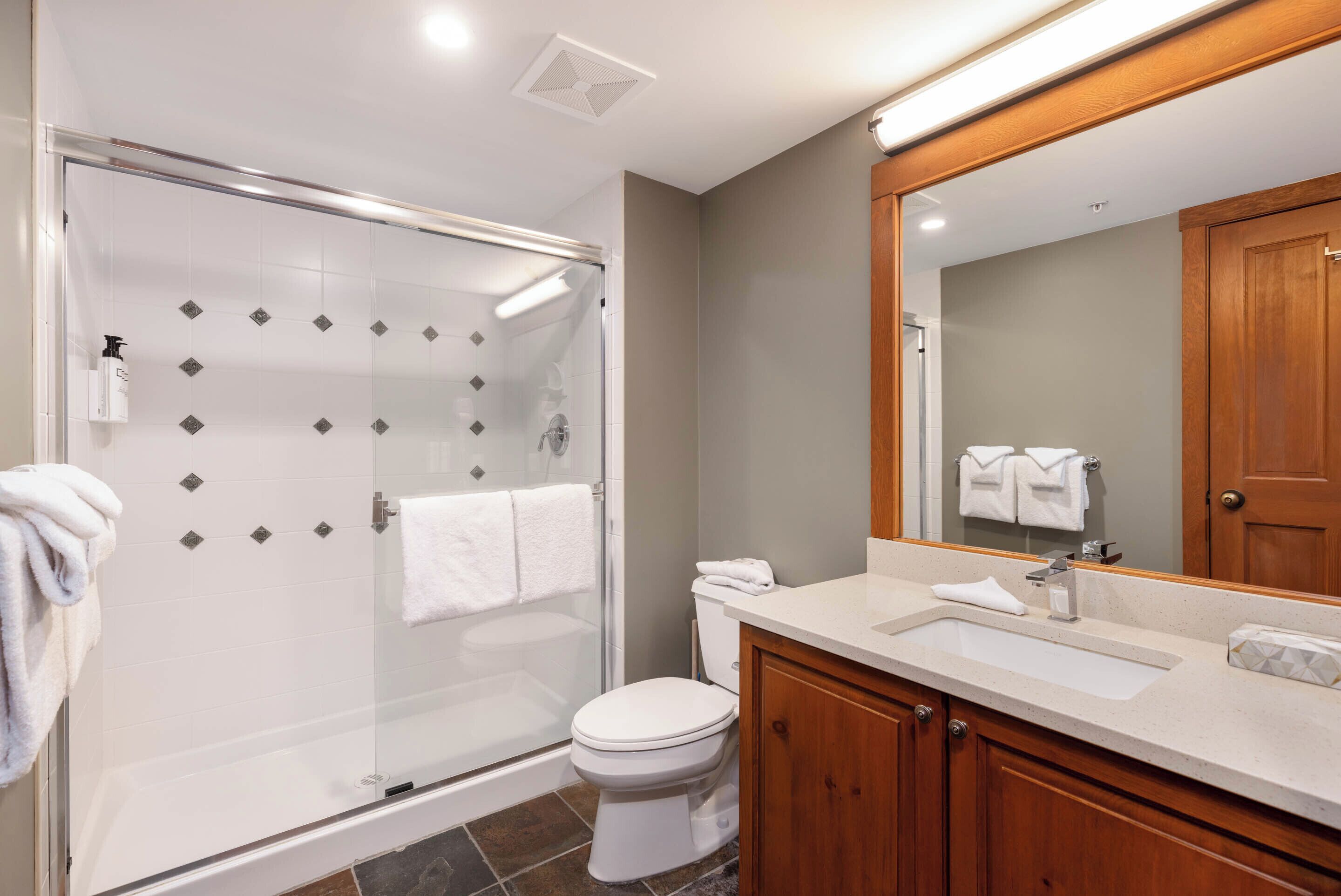 Une salle de bain moderne avec une douche à parois de verre, des toilettes blanches, un meuble-lavabo en bois et un miroir au-dessus du meuble-lavabo.