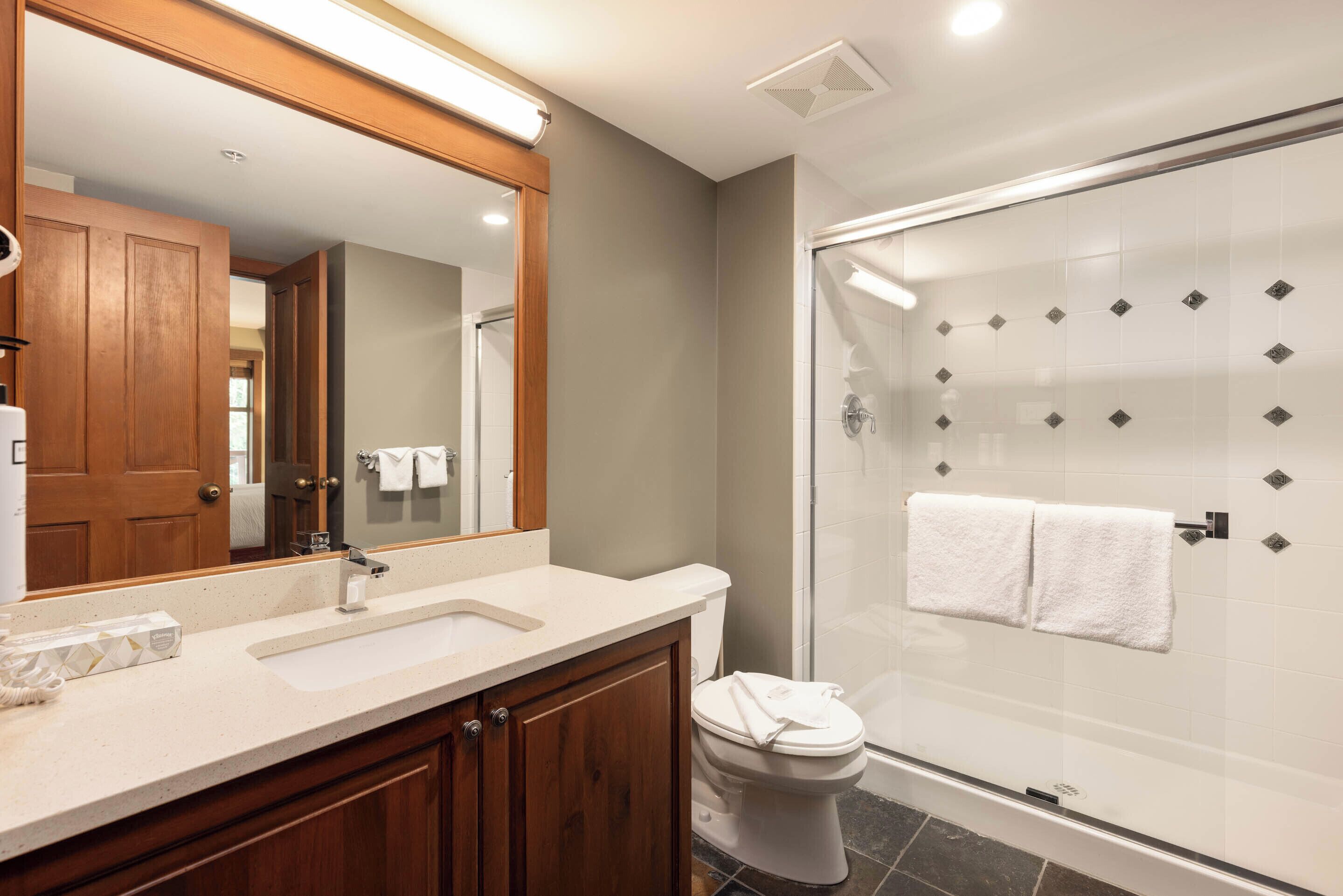 A modern bathroom with a wooden vanity, a large mirror, a glass shower enclosure, and a white toilet.