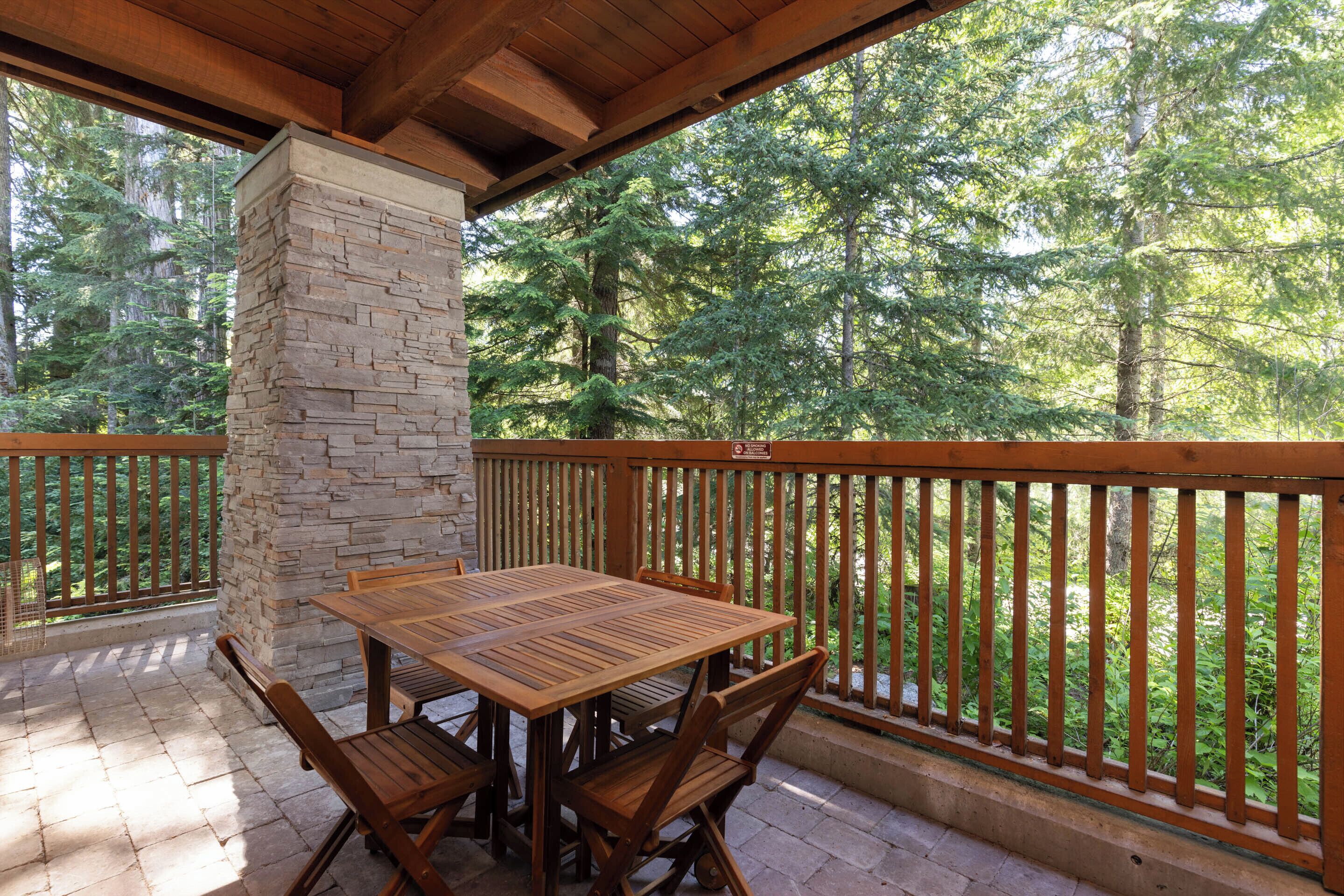 Une terrasse extérieure avec un pilier en pierre, une table et des chaises en bois, entourée d’une balustrade en bois et offrant une vue sur une forêt.