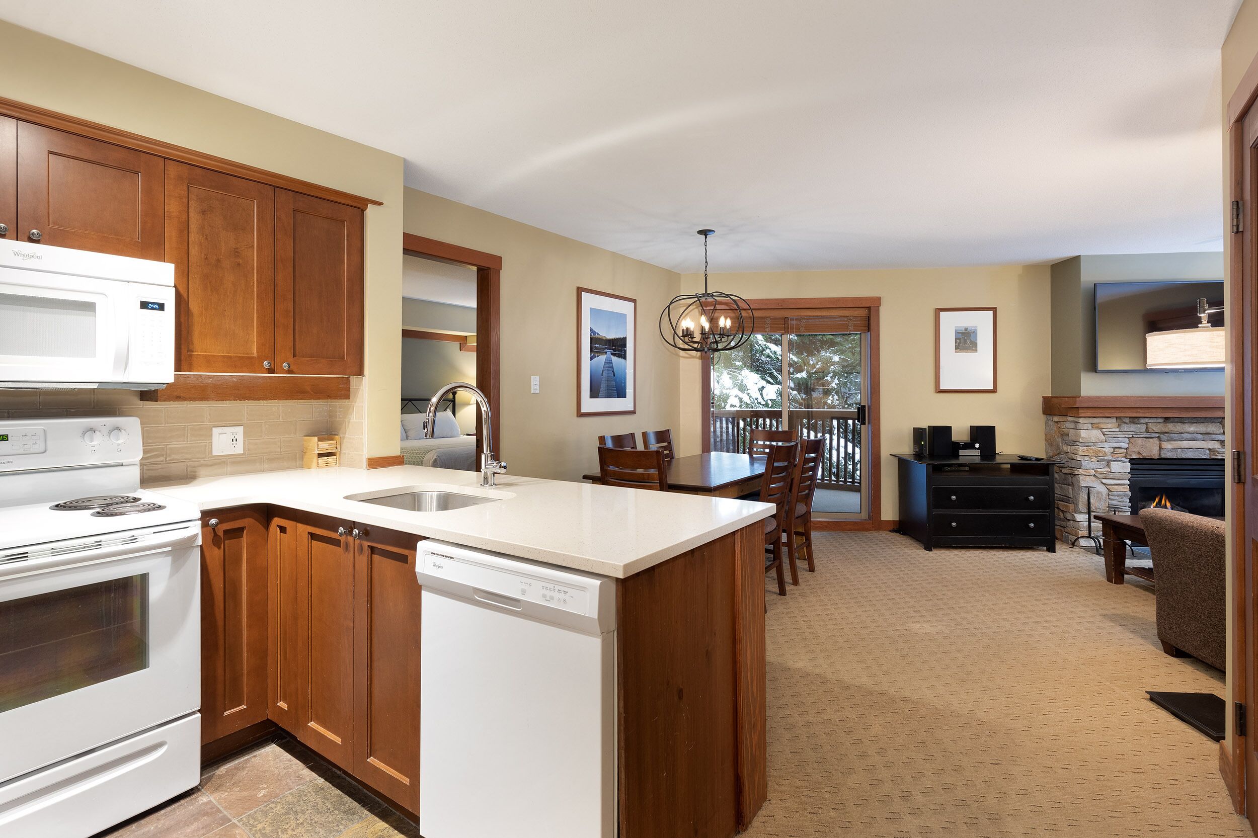 A kitchen with wooden cabinets, a white microwave, and a dishwasher. There is a dining area with a table and chairs, and a fireplace in the background.