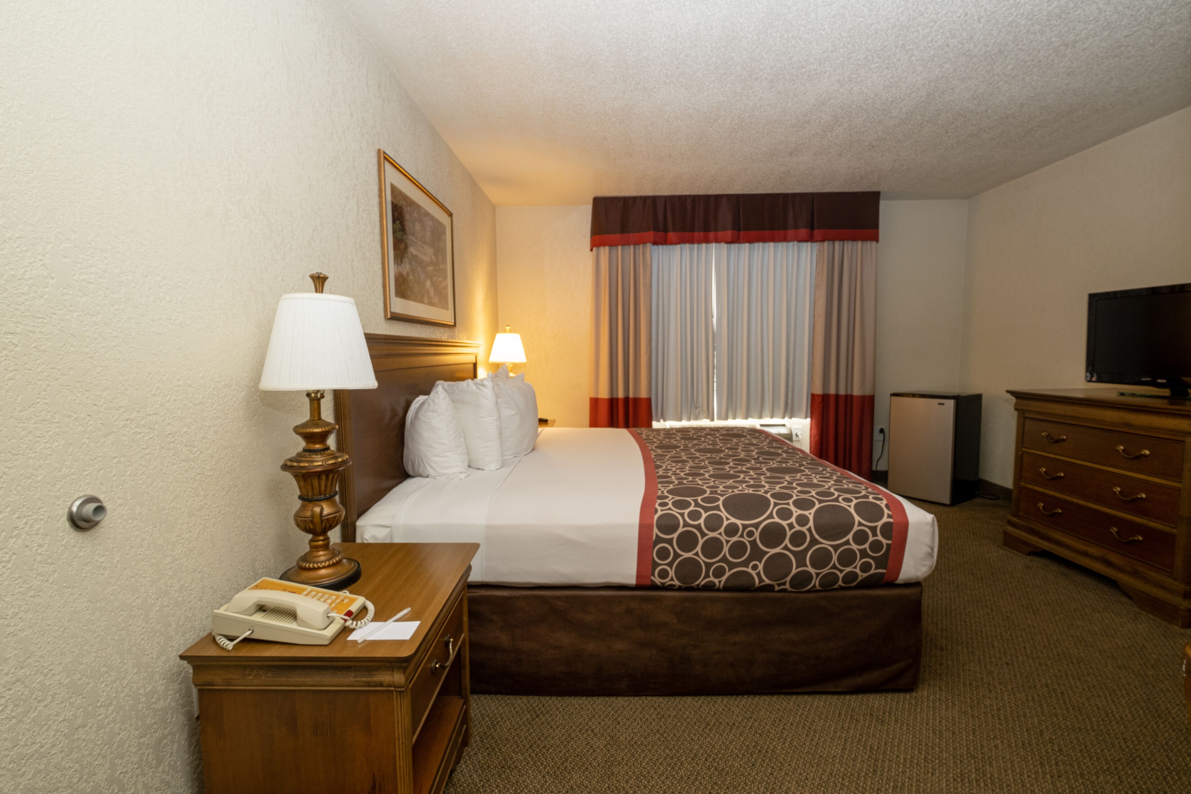 Une chambre d’hôtel comprenant un lit, des lampes de chevet, une commode, un téléviseur et un bureau avec un téléphone et des documents.