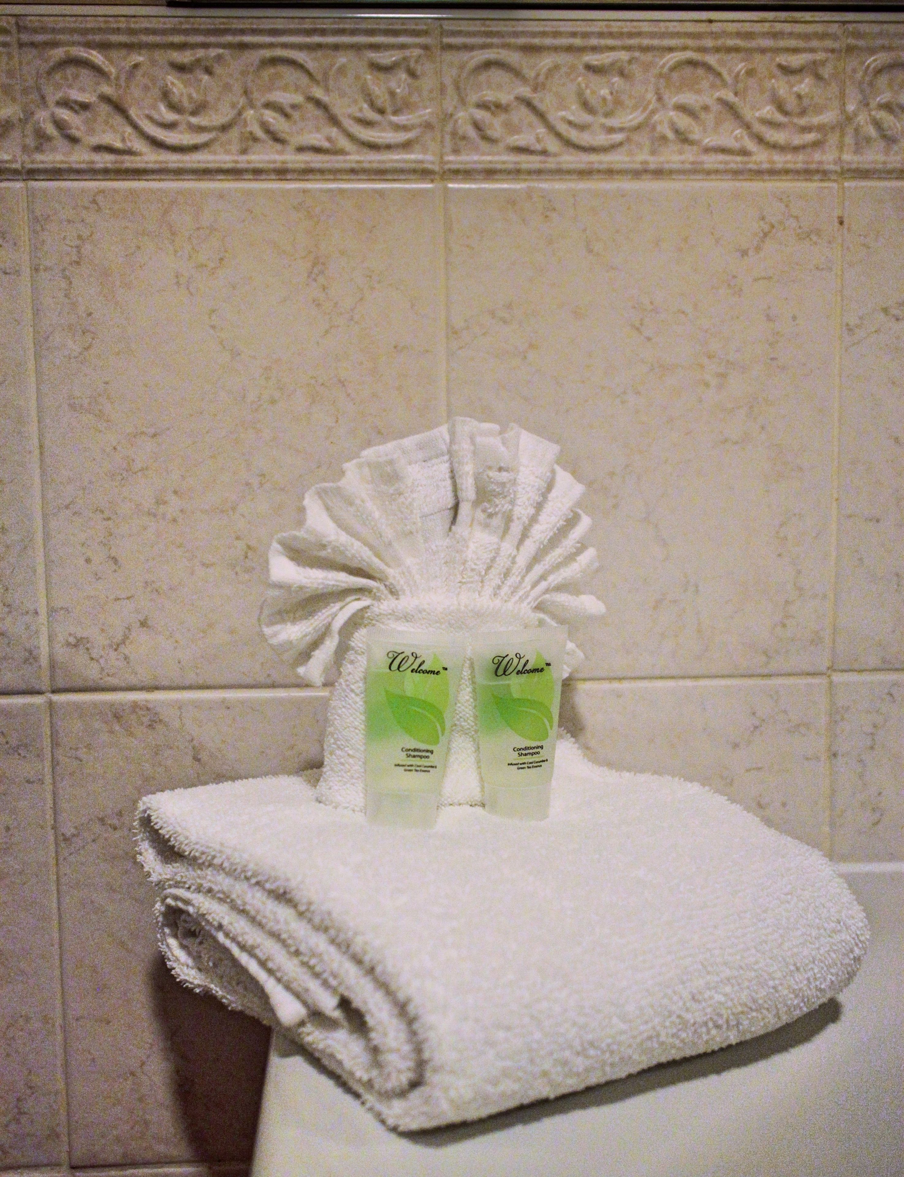 A bathroom with a white towel folded on a white surface, two bottles of 'Welcome' conditioning shampoo, and a tiled wall with a decorative border.