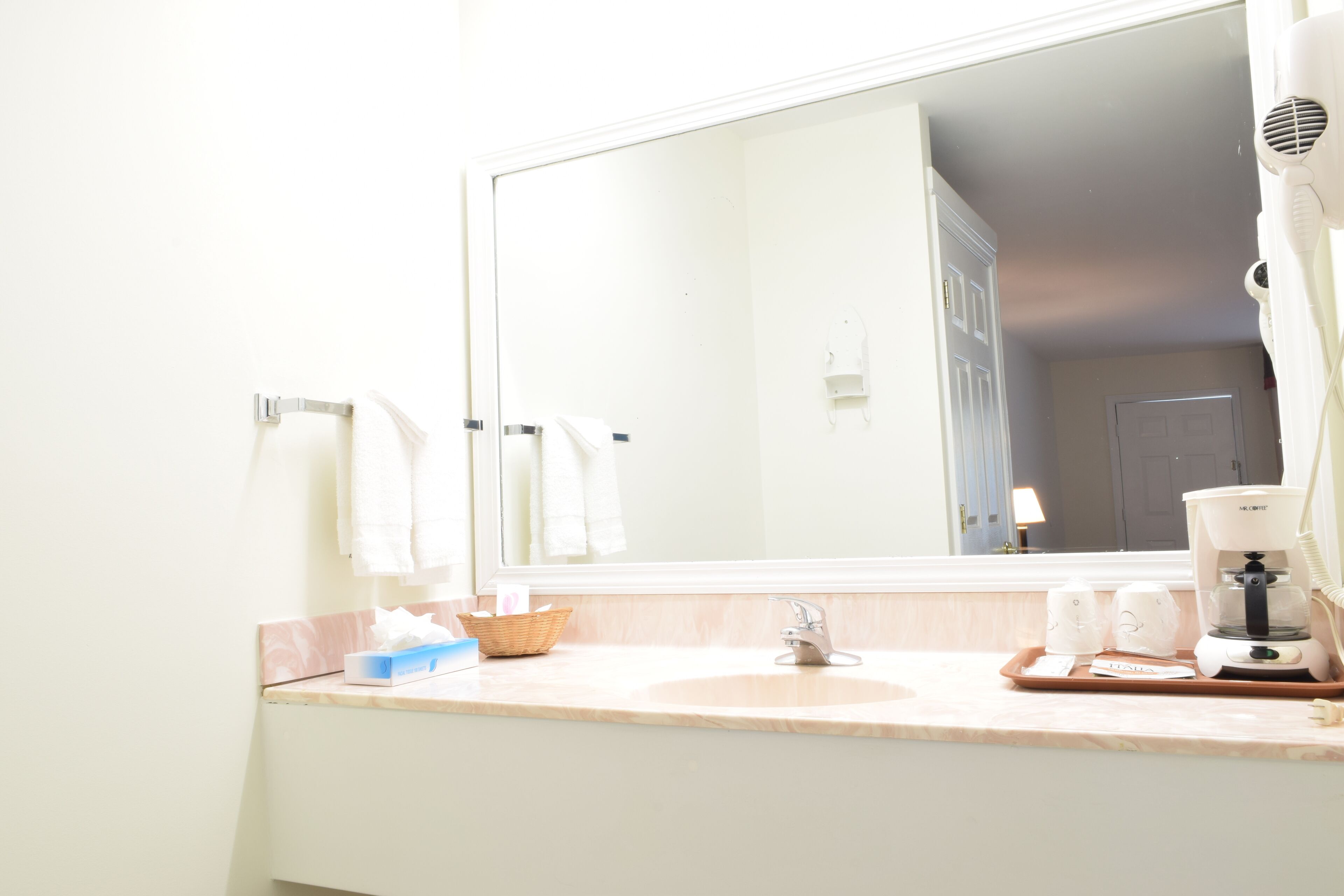 A bathroom with a large mirror, a sink, a coffee maker, and a basket of toiletries.