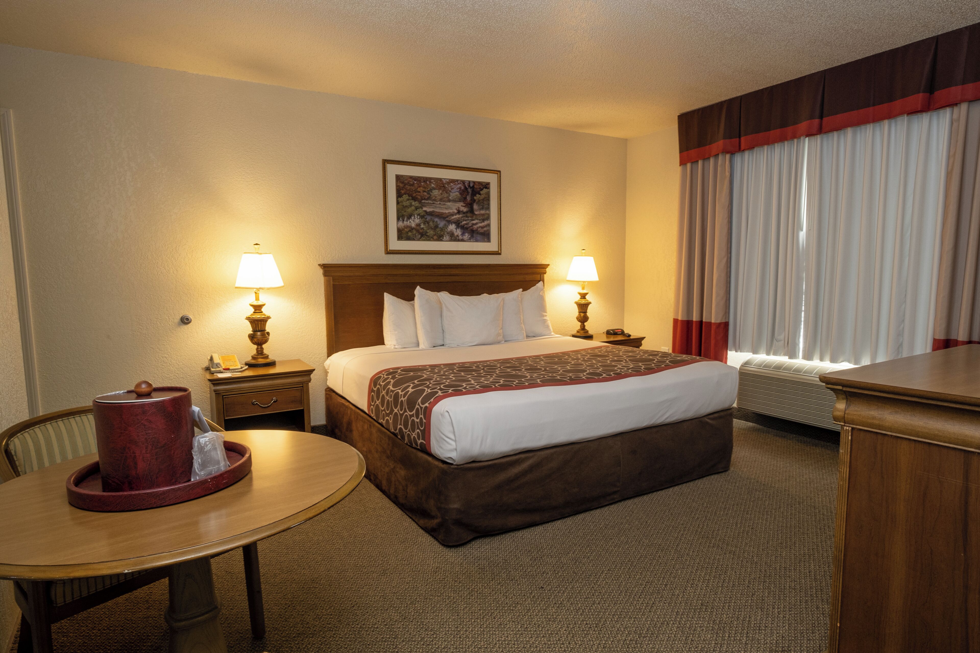 A hotel room with a bed, two bedside lamps, a table with a red object, a chair, a painting on the wall, and a window with curtains.