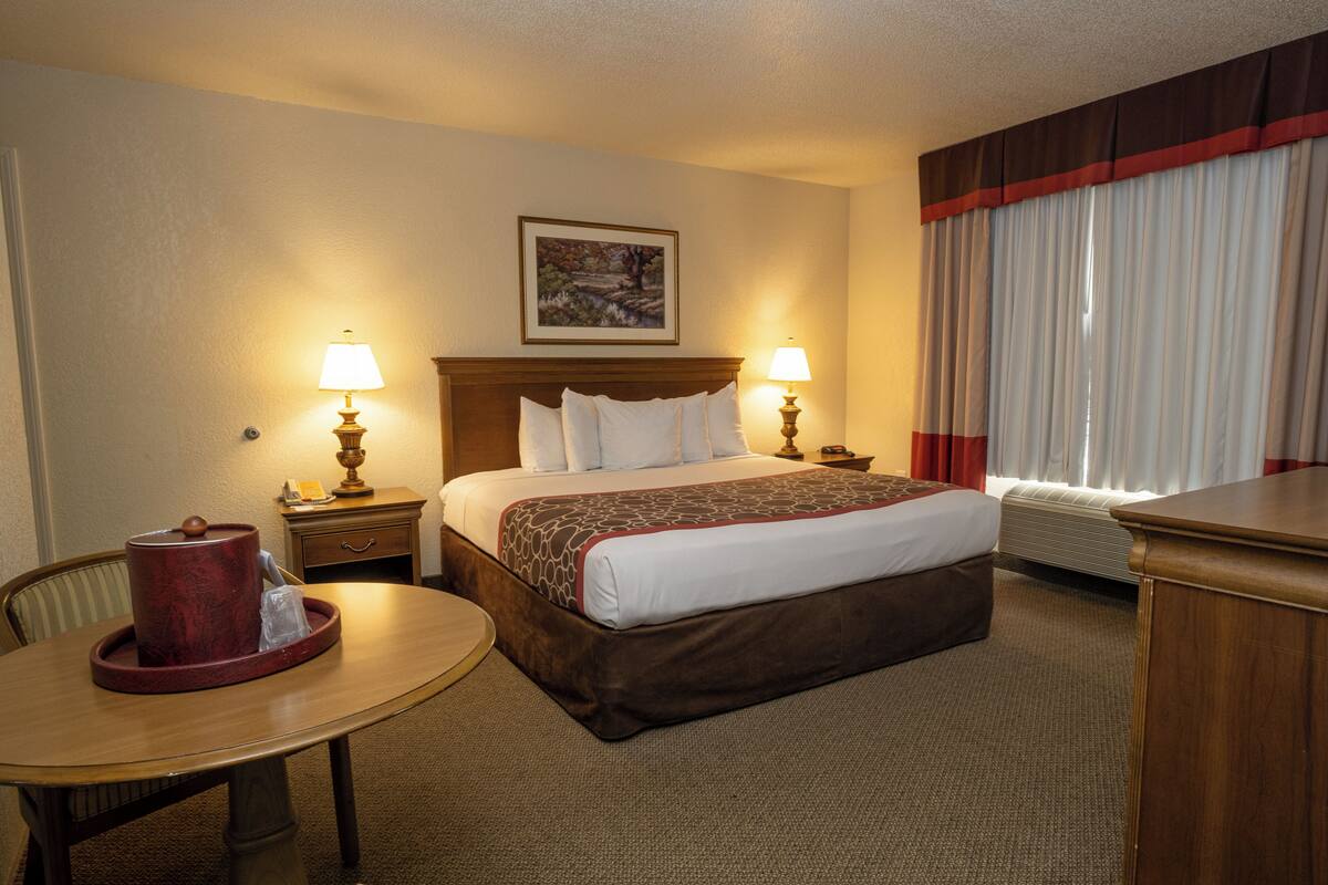 Habitación de hotel con cama, dos lámparas de noche, una mesa con un objeto rojo, una silla, un cuadro en la pared y una ventana con cortinas.