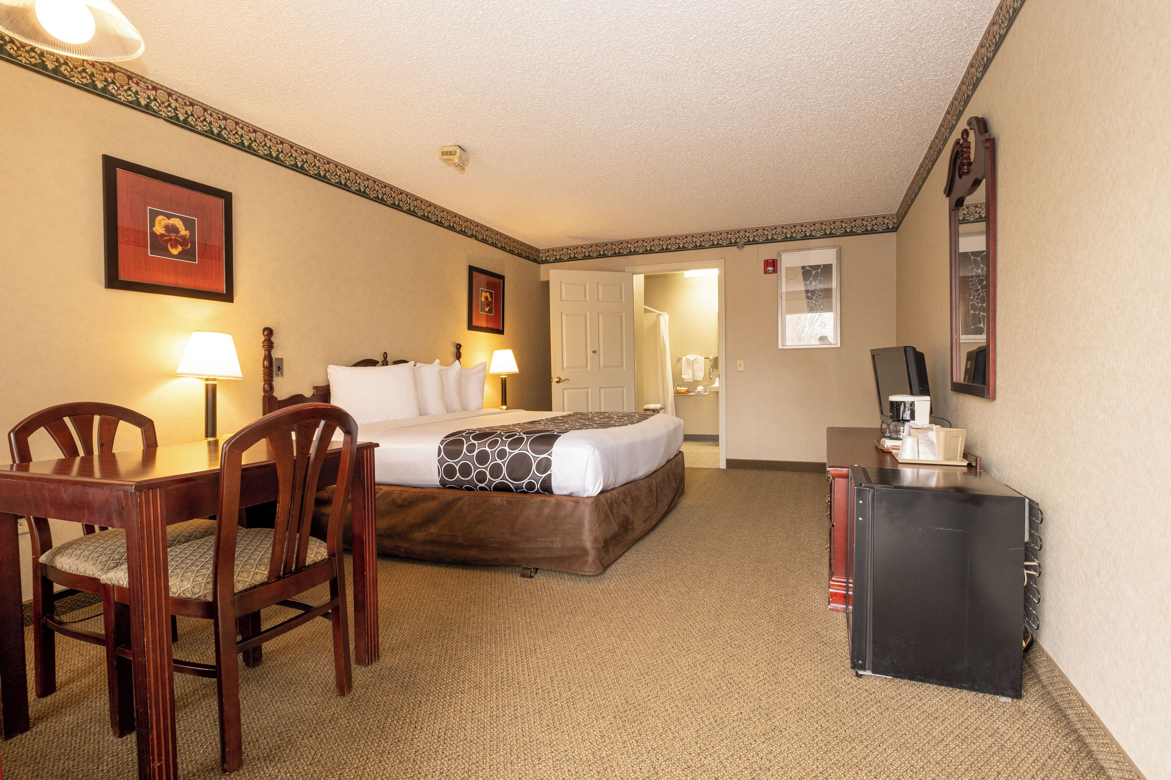 Quarto de hotel com cama, escrivaninha, cadeira, cafeteira e porta para um banheiro.