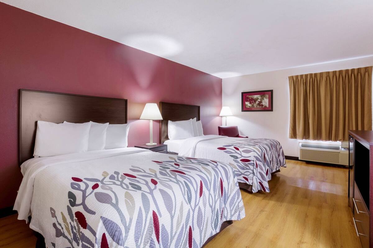 A hotel room with two beds, a window with curtains, a wall-mounted air conditioner, and a framed picture on the wall.