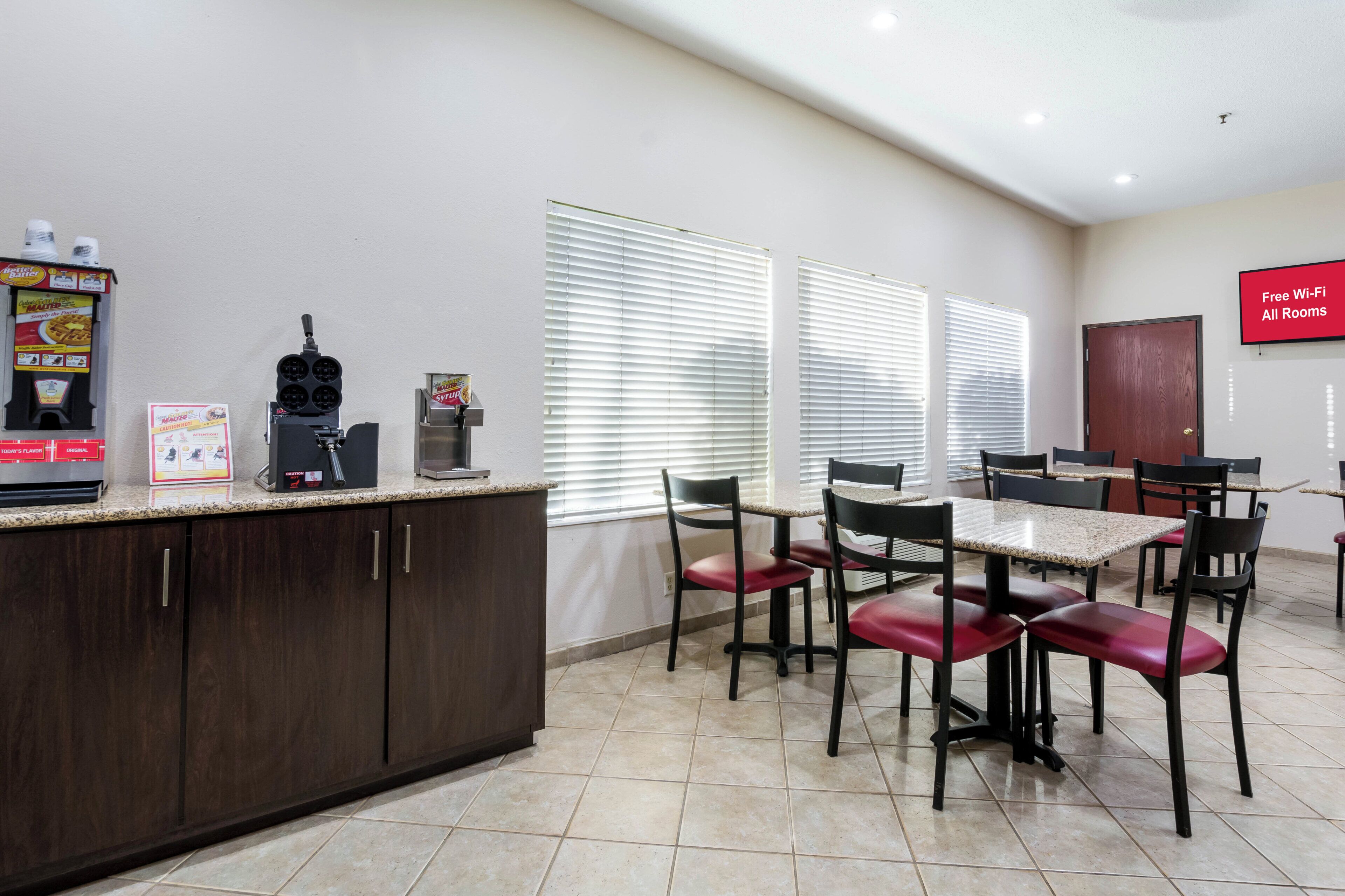 A clean, modern kitchen area with a coffee machine, a waffle maker, and a microwave. There are several chairs and a table, suggesting a dining area.