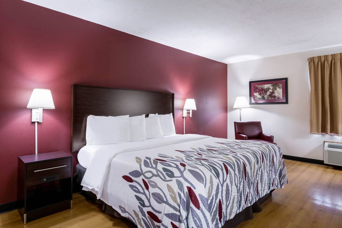 A hotel room with a large bed, two bedside tables, a chair, a desk, and a wall-mounted air conditioner.