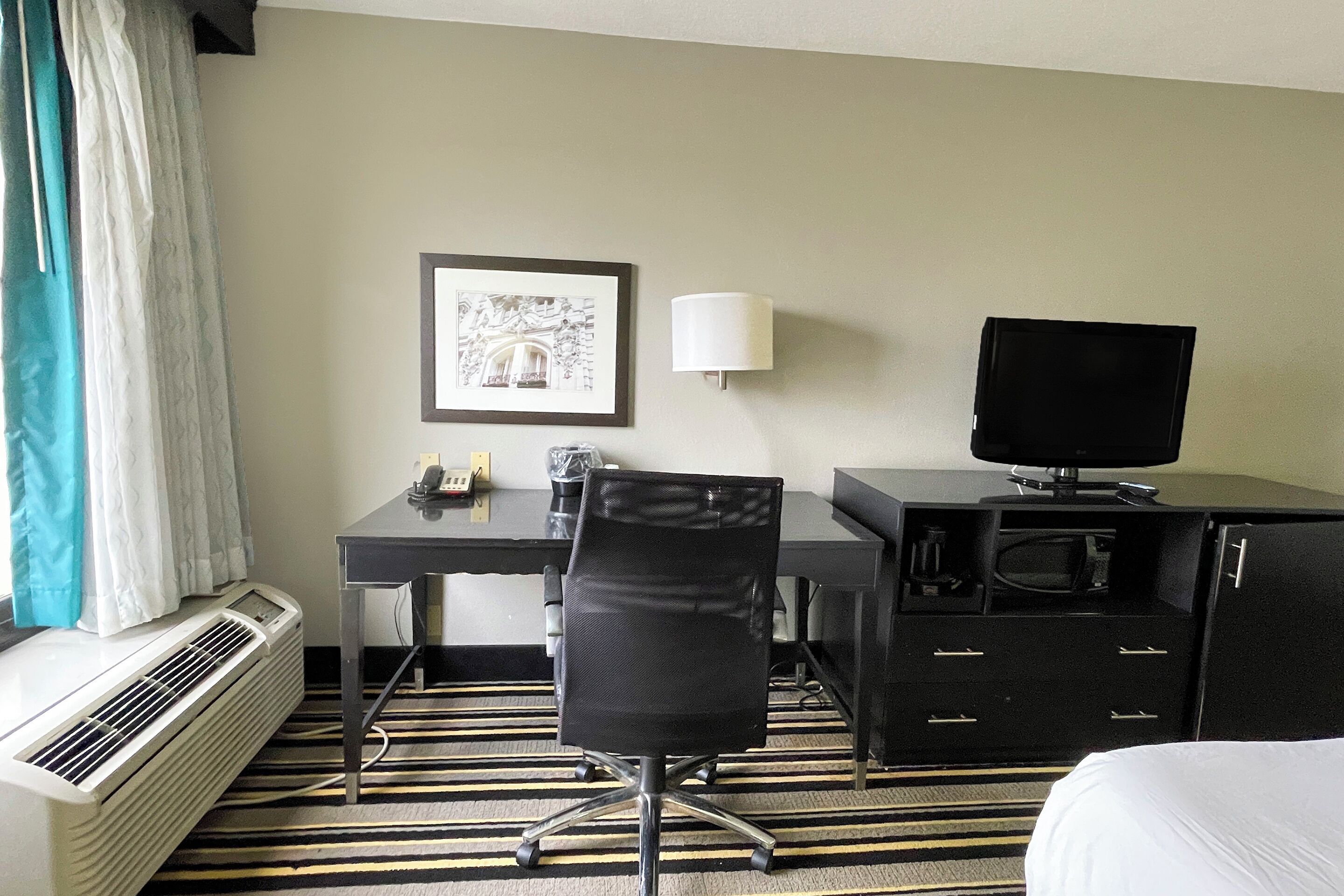 A hotel room with a bed, a desk with a computer, a chair, a lamp, a television, and a heater.