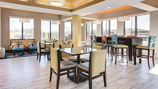Un amplio comedor de hotel con varias mesas y sillas, ventanas grandes que ofrecen vistas del paisaje urbano y un diseño de techo moderno.