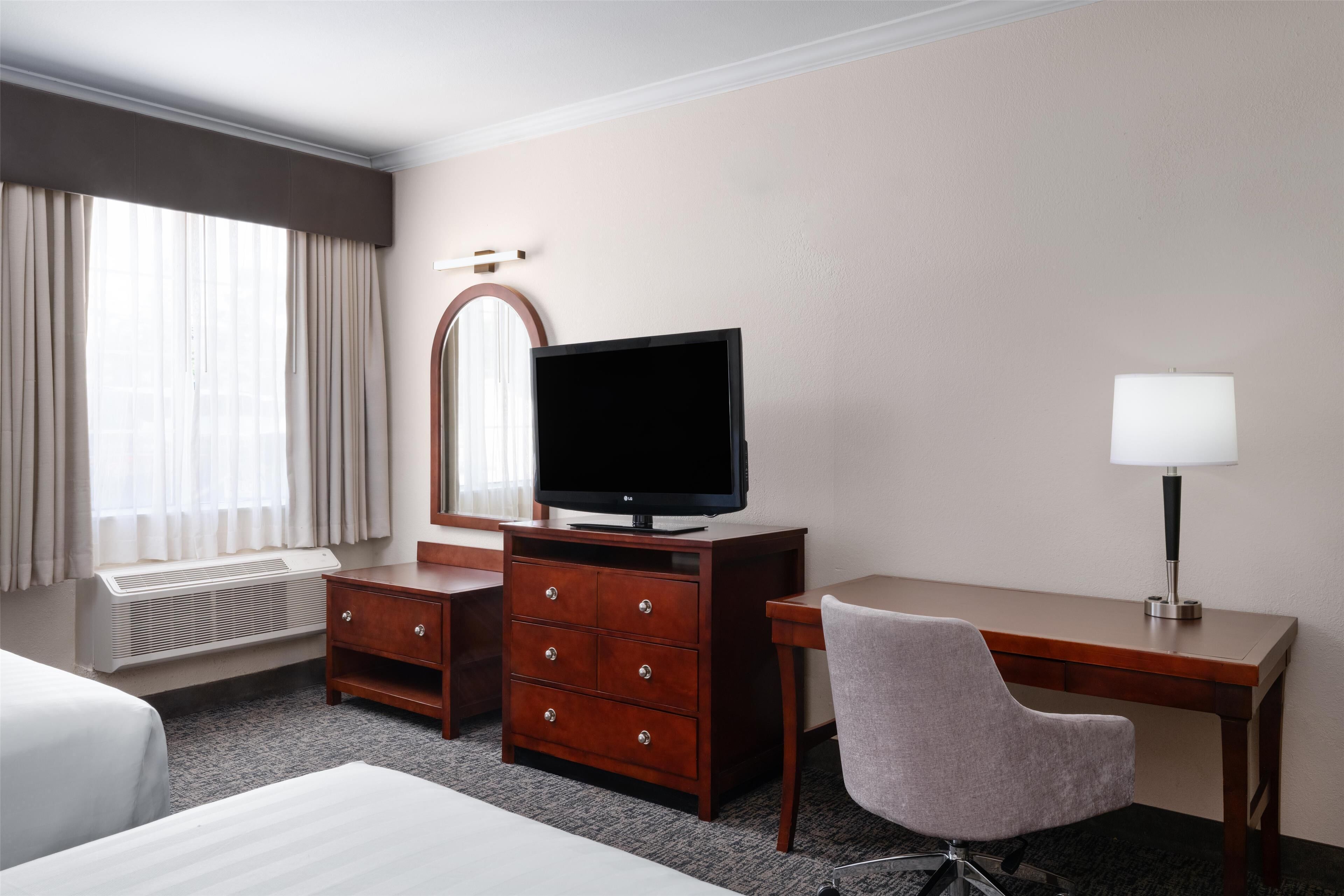 A hotel room with a bed, a desk with a chair, a television, a lamp, and a mirror.