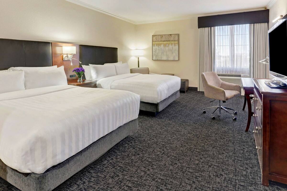 A hotel room with two beds, a desk, a chair, a TV, and a window with curtains.