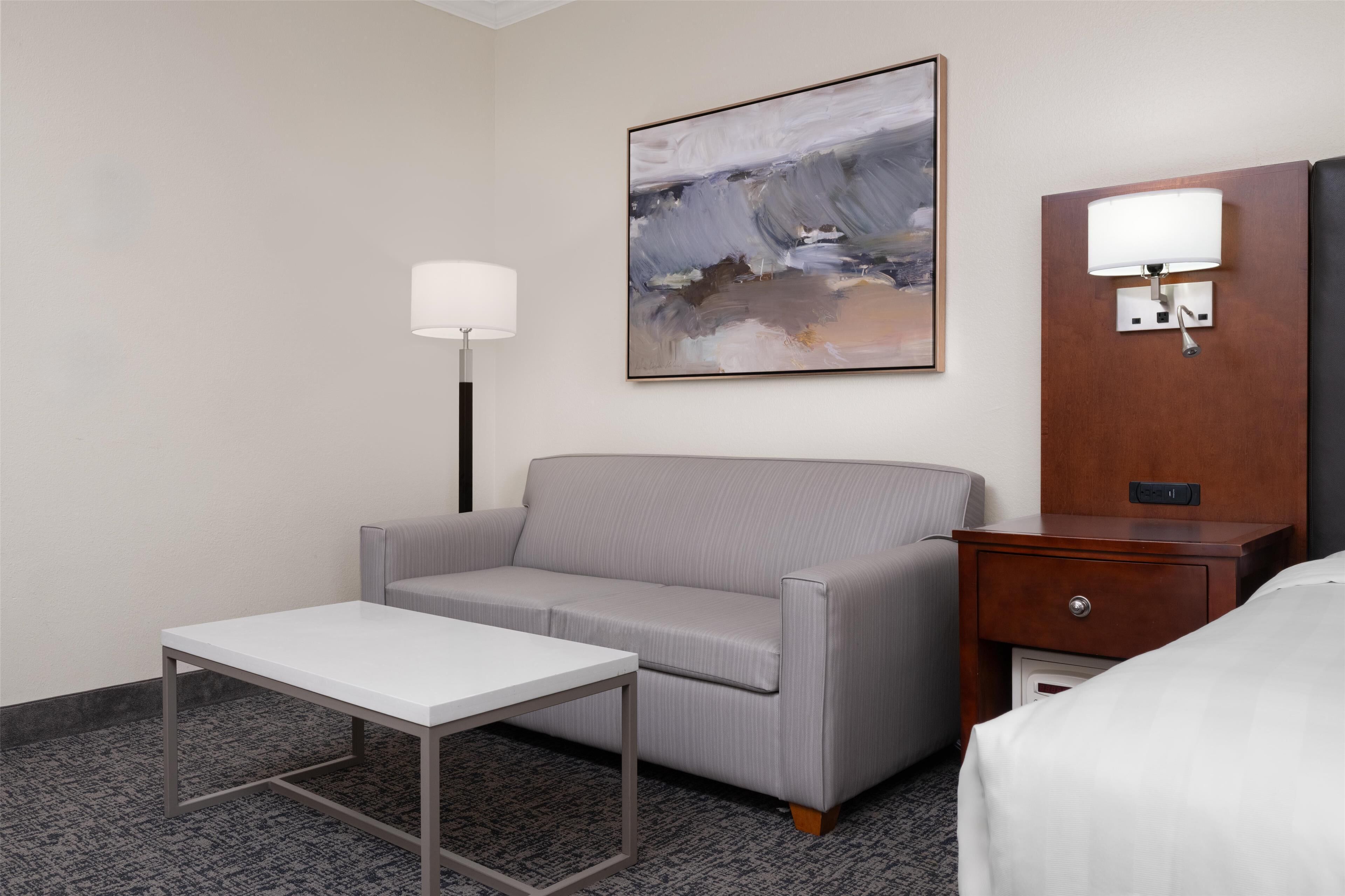 A hotel room with a grey sofa, a small white table, a lamp, a wooden nightstand with a lamp, and a painting on the wall.