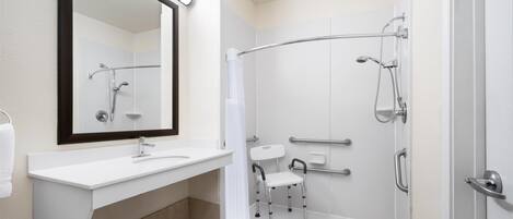 A modern bathroom with a white sink, a large mirror, a shower with a curved glass door, and a wheelchair-accessible bench.
