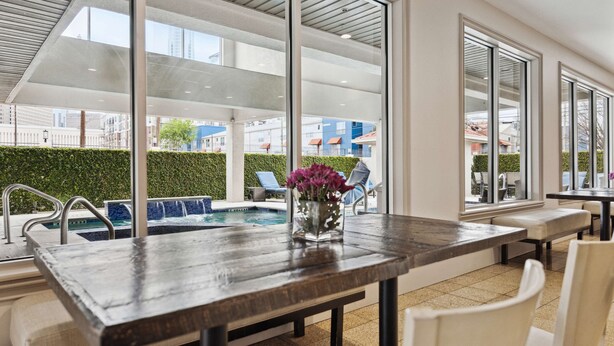 Area piscina interna moderna con tavolo e sedie in legno, un vaso con fiori e vista sugli edifici esterni.