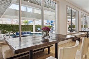 Un área de piscina interior moderna con una mesa y sillas de madera, un jarrón con flores y vistas a edificios exteriores.