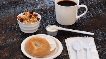 Una mesa de madera con desayuno: bagel, granola, yogur, café y cubiertos.