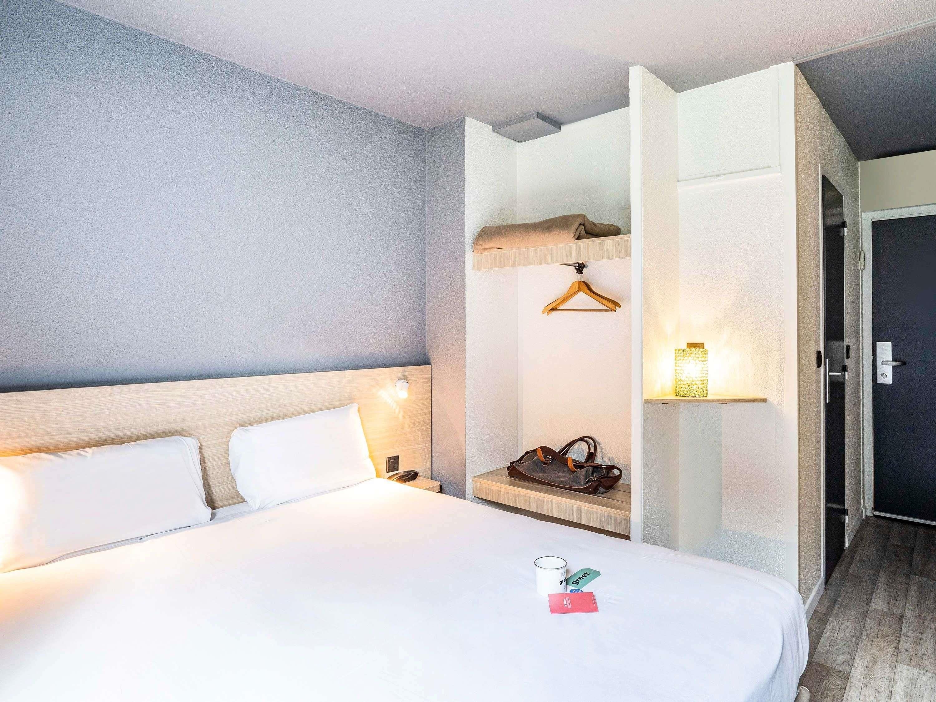 A hotel room with a bed, a bedside table, a hanging coat rack, and a door leading to another room.