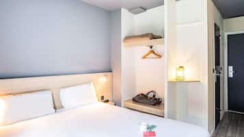 A hotel room with a bed, a bedside table, a hanging coat rack, and a door leading to another room.