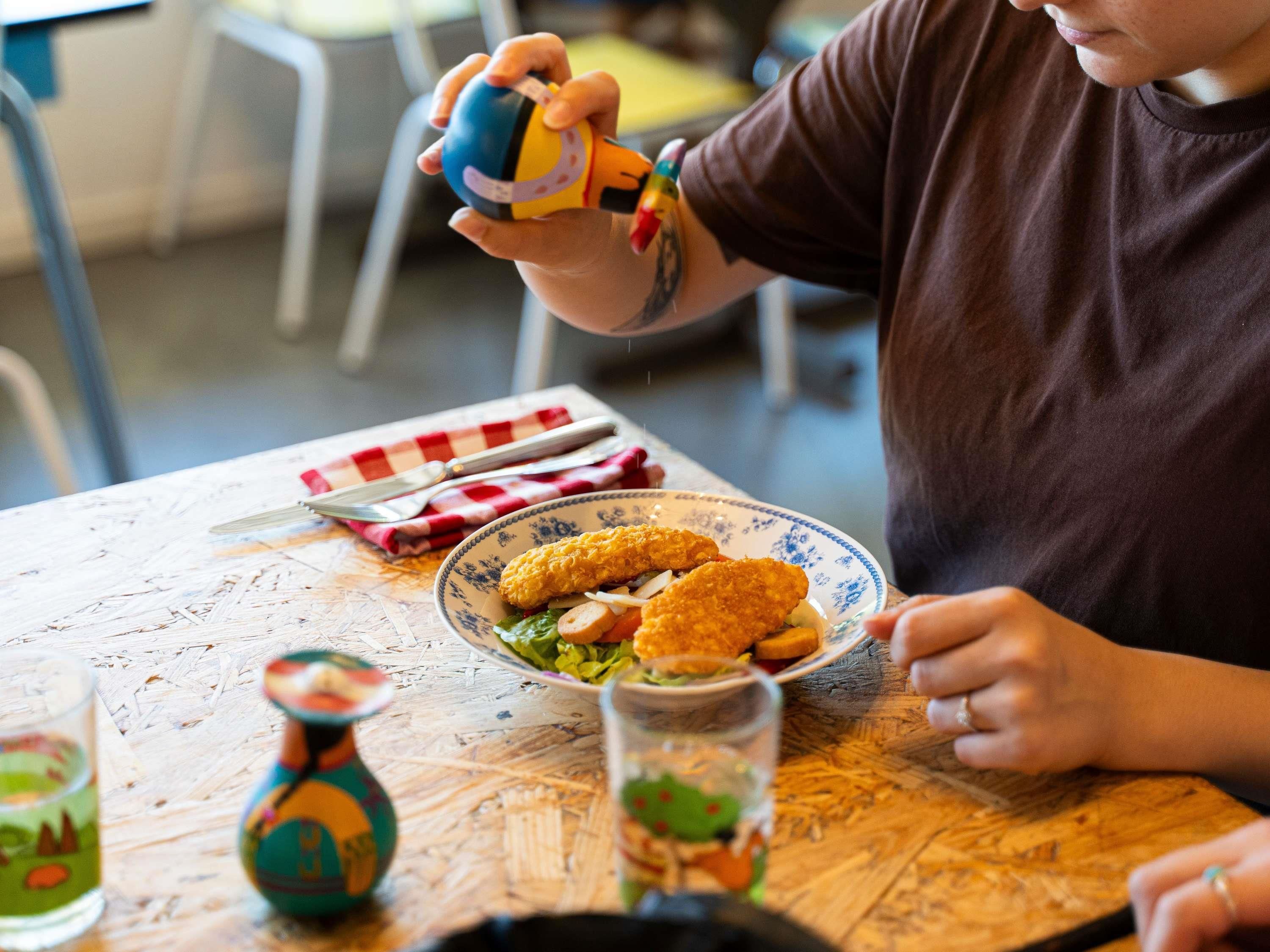 Una persona sostiene un juguete colorido sobre una mesa de madera con un plato de comida, un vaso y una botella decorativa.