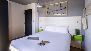 Una cama bien hecha con un ordenador portátil y una revista, una obra de arte abstracto colgada en la pared, y una mesita de noche verde con una lámpara.