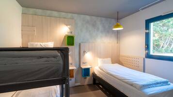 Una habitación con literas, una cama individual, una ventana con vistas a árboles, una lámpara colgante amarilla y un estante verde.