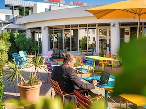 Iemand werkt op een laptop in een openlucht café met kleurrijke stoelen en tafels, met een gebouw met grote ramen op de achtergrond.