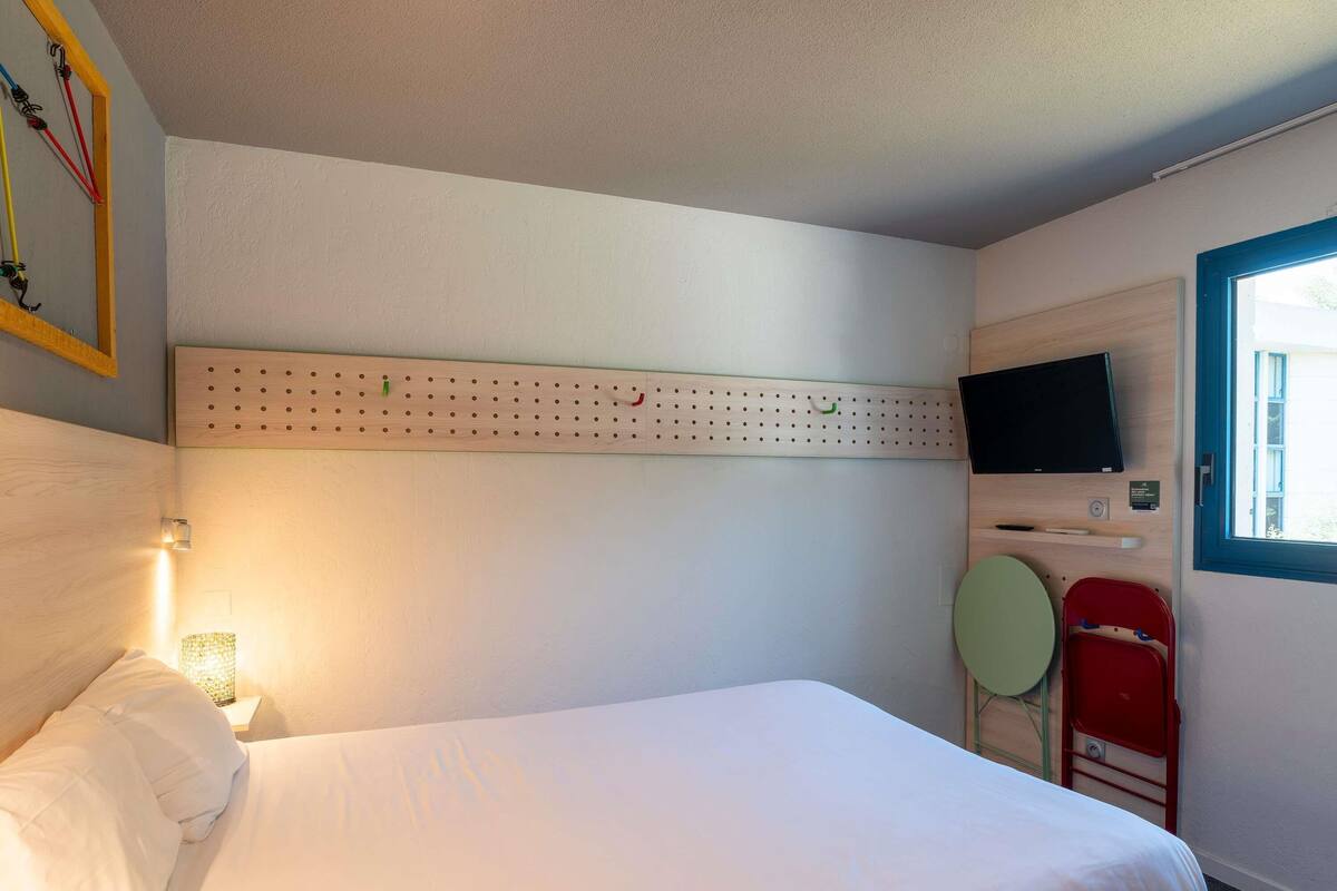 Una habitación de hotel con una cama, un televisor montado, un tablero de corcho y una ventana.