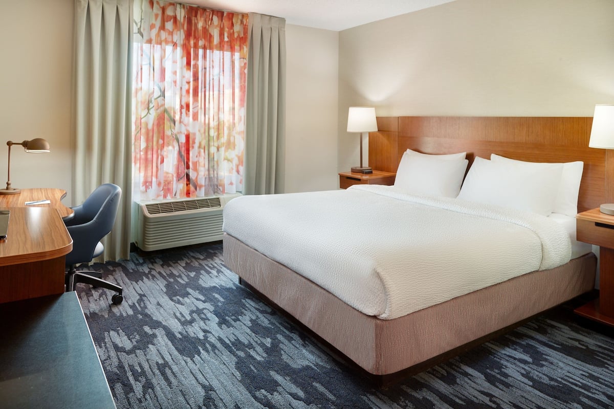 Una habitación de hotel con una cama grande, un escritorio con una silla, una lámpara de escritorio y una ventana con cortinas.