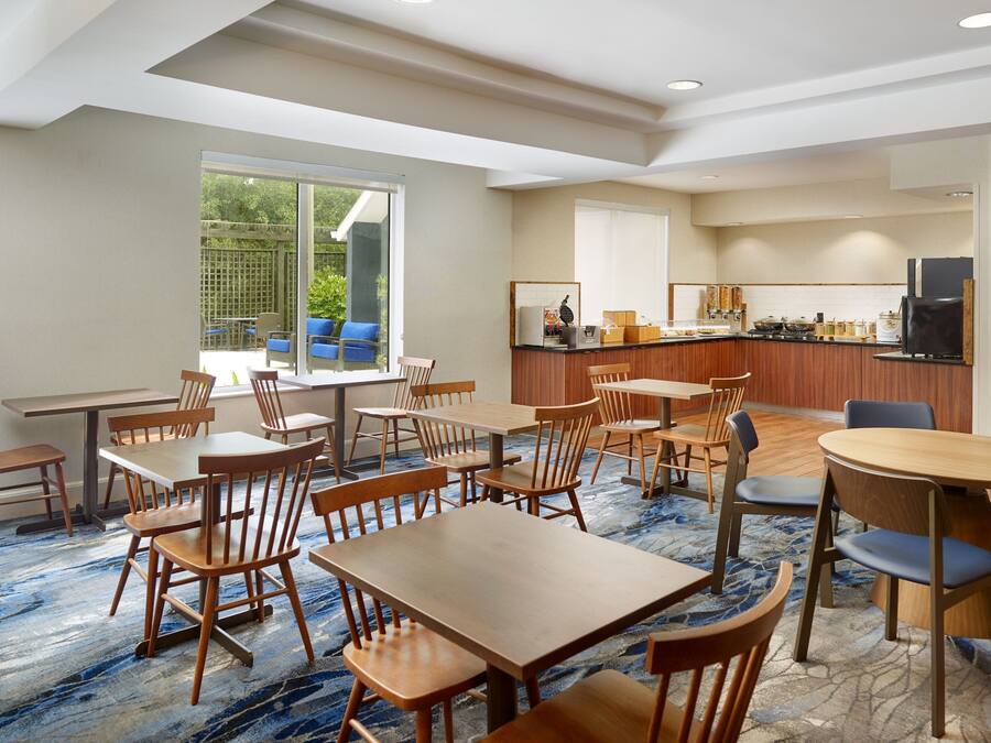 Un comedor de hotel con varias mesas y sillas, una cocina al fondo y un ventanal con vistas al exterior.