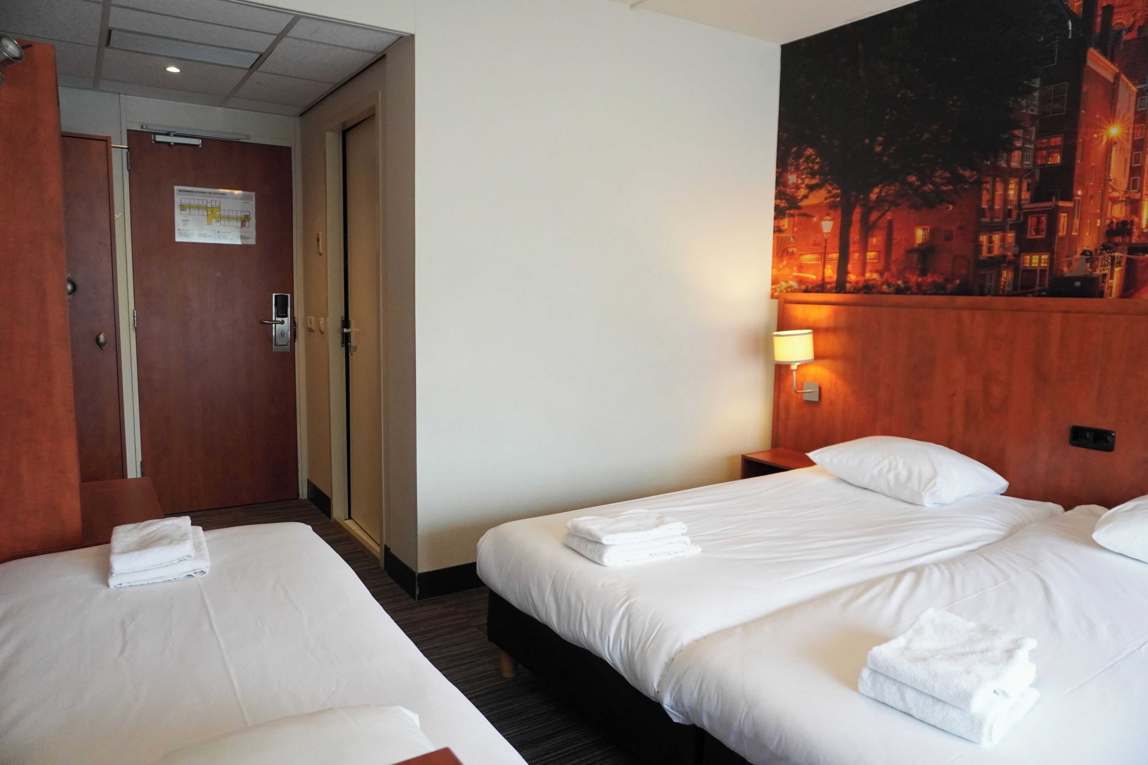A hotel room with two beds, a wooden headboard, a night lamp, and a cityscape painting on the wall.