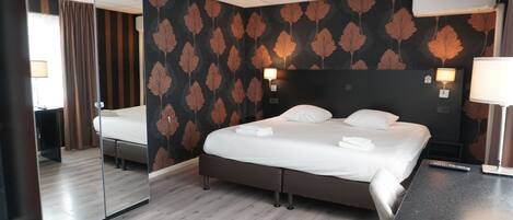 A hotel room with a bed, a mirror, a bedside table, a lamp, and a wall with a leaf pattern.