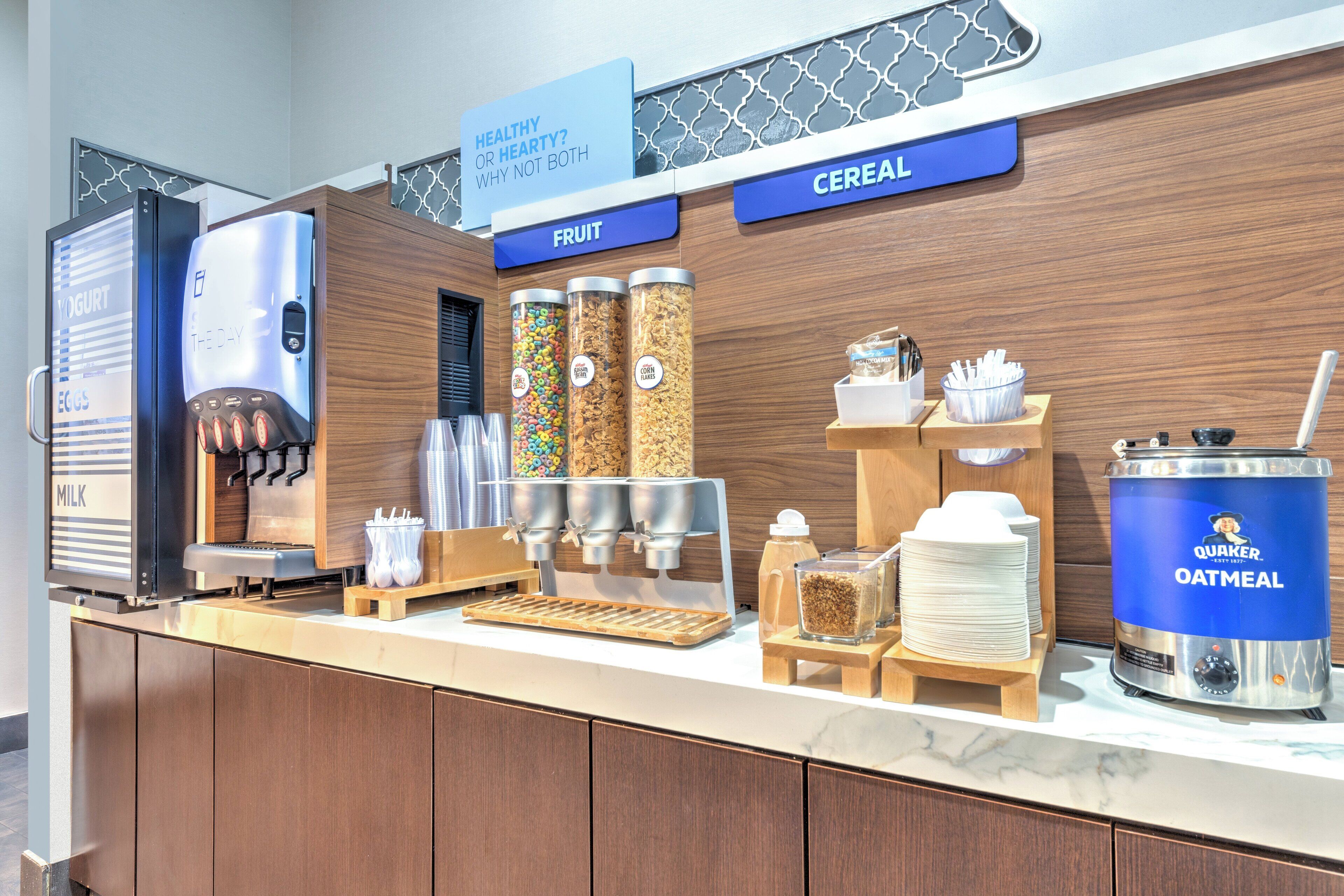 En selvbetjenings morgenmadsstation med yoghurt, cornflakes og frugtautomater, samt en maskine til mælk og en havregrydsdispenser.