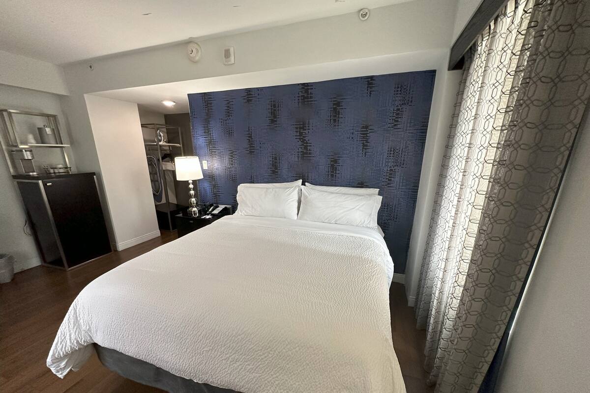 A hotel room with a large bed, a desk with a lamp, a small refrigerator, and a bathroom visible in the background.