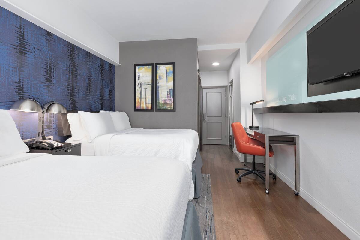A hotel room with two beds, a desk, a chair, a TV, and artwork on the wall.