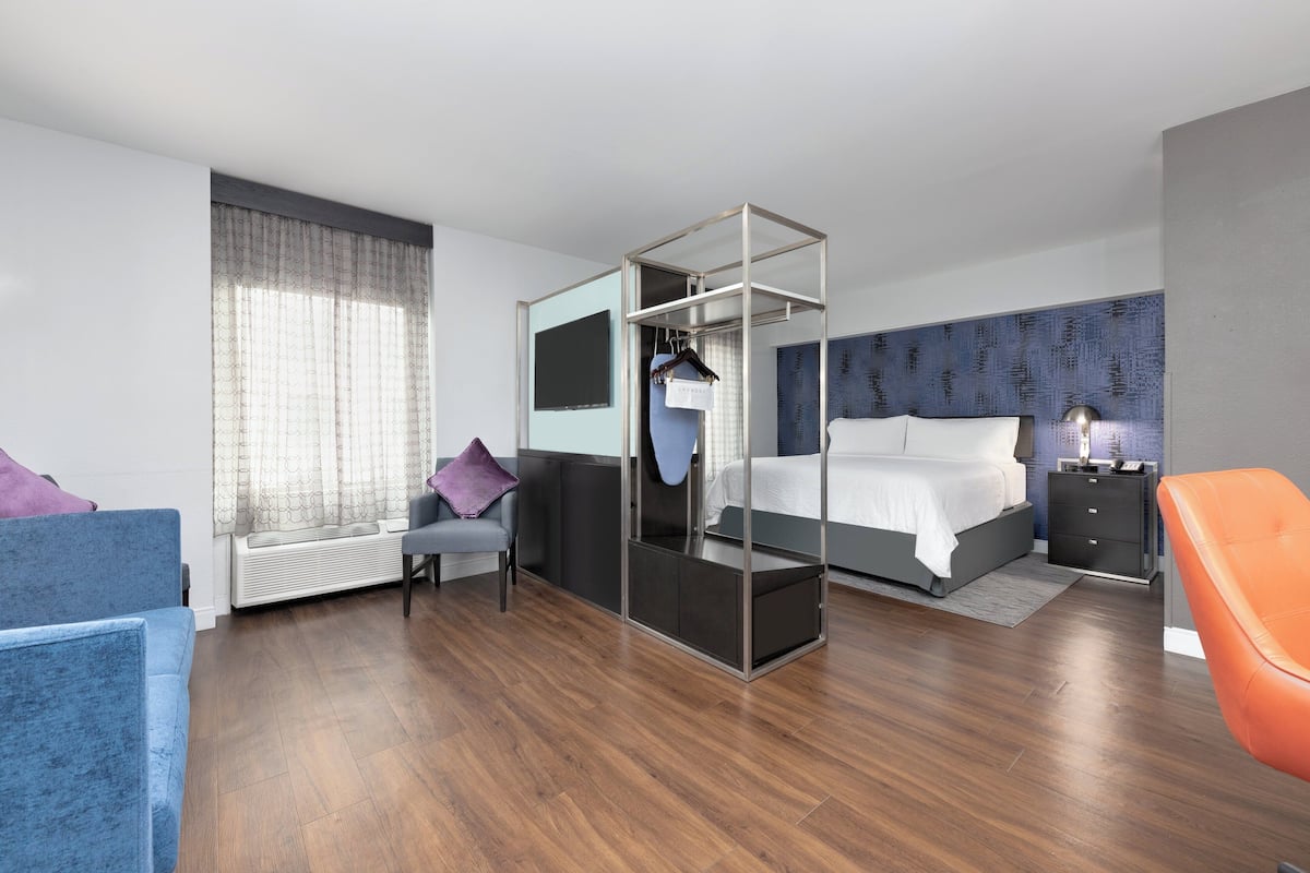 A modern hotel room with a bed, a seating area with a chair, a small table, and a wardrobe.