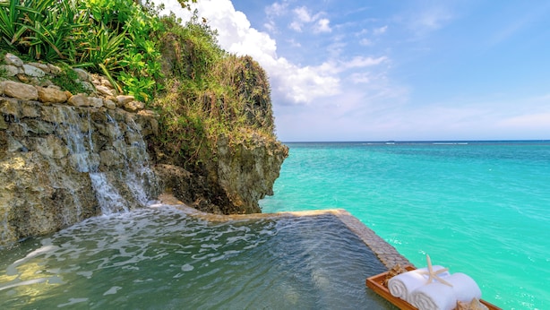 A clear view of a pool with a wooden edge, a wooden tray with towels and a seashell, and a natural rocky cliff with a small waterfall.