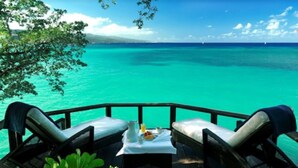 A deck with two lounge chairs and a table, overlooking turquoise waters and a clear sky.