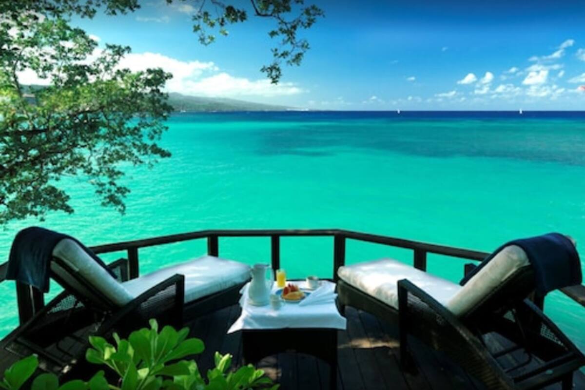 A deck with two lounge chairs and a table, overlooking turquoise waters and a clear sky.