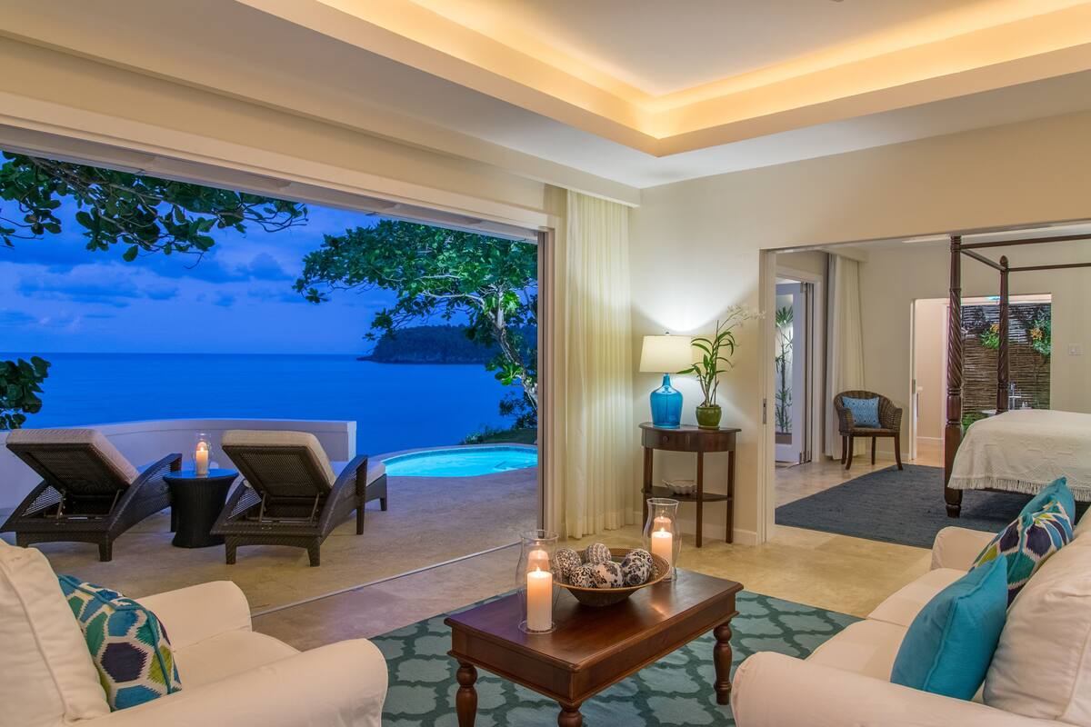 A hotel room with a balcony overlooking a pool and the sea, a bed, a sitting area with a table, and a view of the sea.