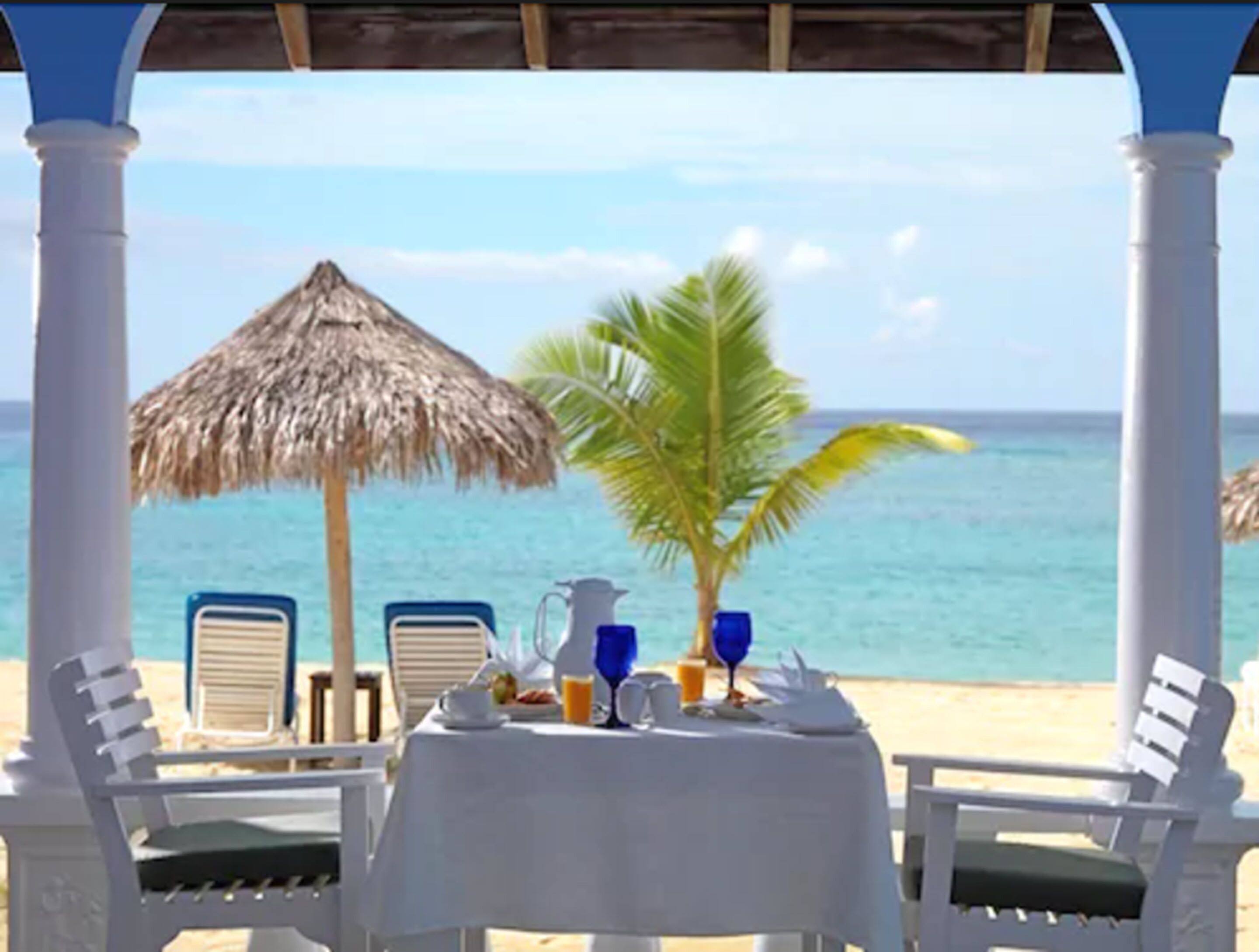 A beachside dining setup with a table set for two, a thatched umbrella, and a palm tree.
