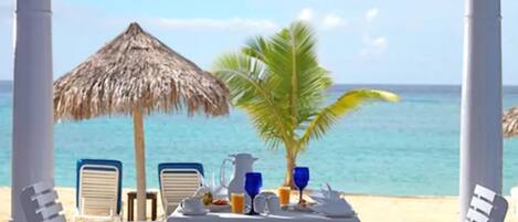 Un coin repas aménagé sur la plage, avec une table dressée pour deux personnes, un parasol en feuilles de palmier et un palmier.